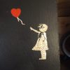 Gold-etched silhouette of a young girl reaching toward a red heart-shaped balloon on a black slate plaque, conveying a sense of longing and innocence.