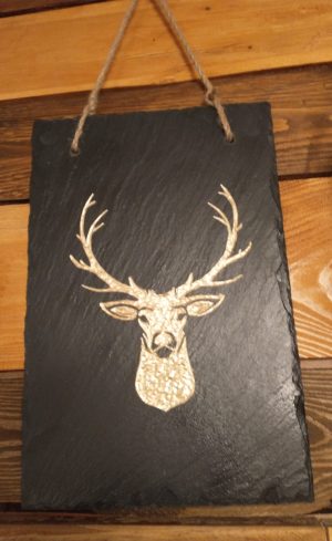 A rustic slate wall hanging with a gold-etched deer head design, featuring detailed antlers and a natural slate texture. The plaque is suspended by a thick jute rope, set against a warm, wooden background, emphasizing the piece's natural, earthy appeal.